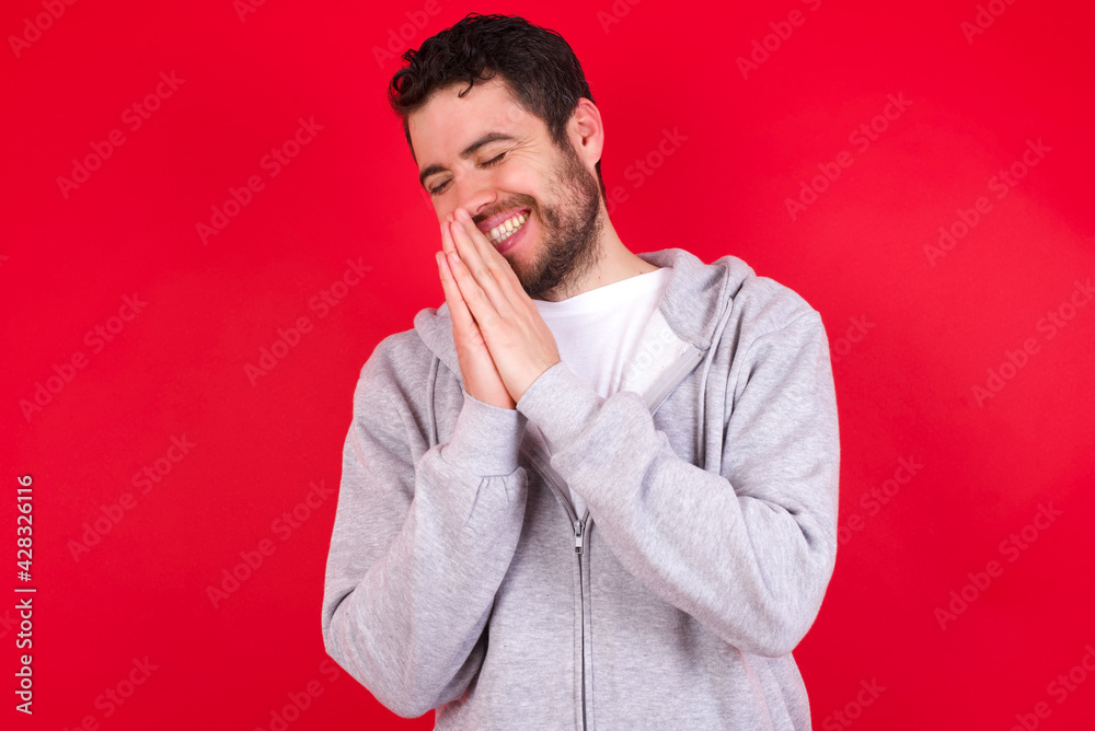 Overjoyed young handsome caucasian man in sports clothes against red wall laughs joyfully and keeps palms pressed together hears something funny