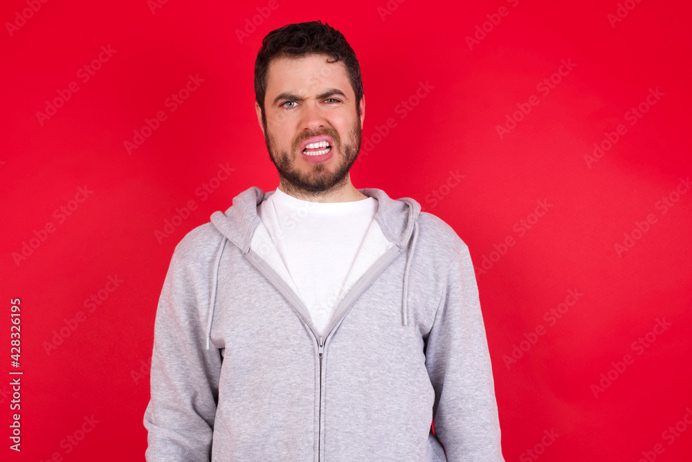 Portrait of dissatisfied young handsome caucasian man in sports clothes against red wall smirks face, purses lips and looks with annoyance at camera, discontent hearing something unpleasant Portrait of dissatisfied young handsome caucasian man in sports clothes against red wall smirks face, purses lips and looks with annoyance at camera, discontent hearing something unpleasant