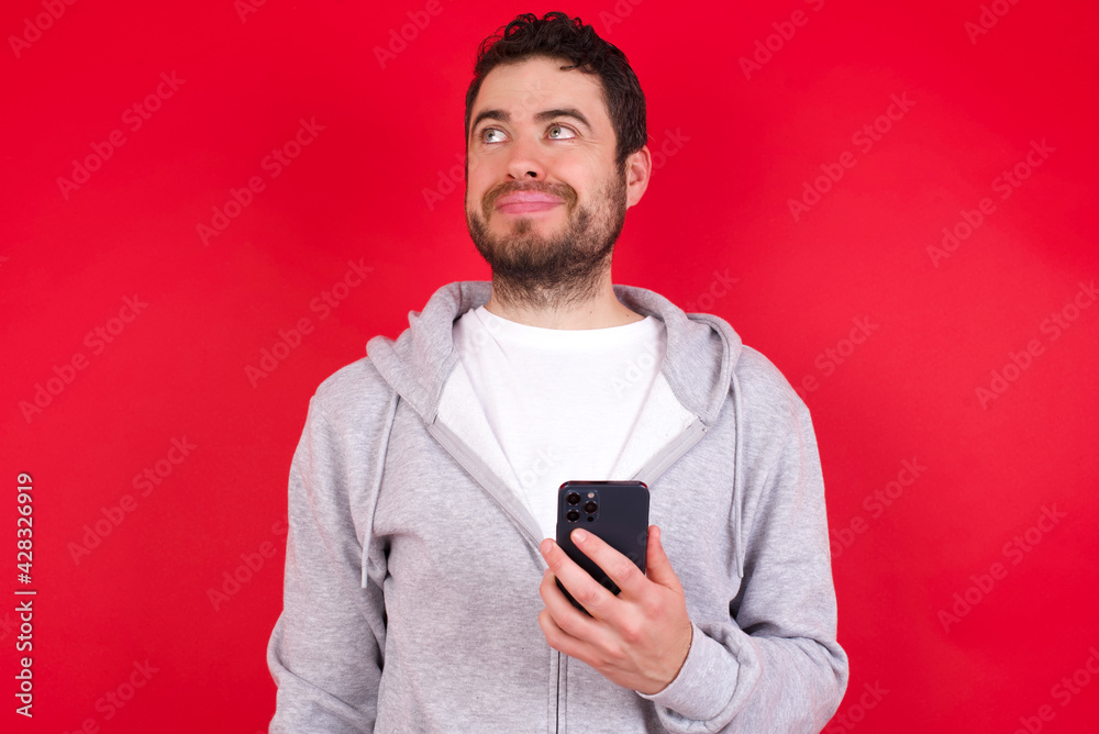 young handsome caucasian man in sports clothes against red wall holds mobile phone uses high speed internet and social networks has online communication. Modern technologies concept young handsome caucasian man in sports clothes against red wall holds mobile phone uses high speed internet and social networks has online communication. Modern technologies concept
