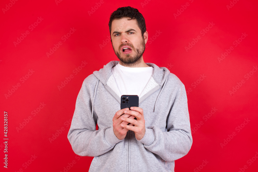 Photo of astonished crazy young handsome caucasian man in sports clothes against red background hold smartphone dislike feedback concept Photo of astonished crazy young handsome caucasian man in sports clothes against red background hold smartphone dislike feedback concept
