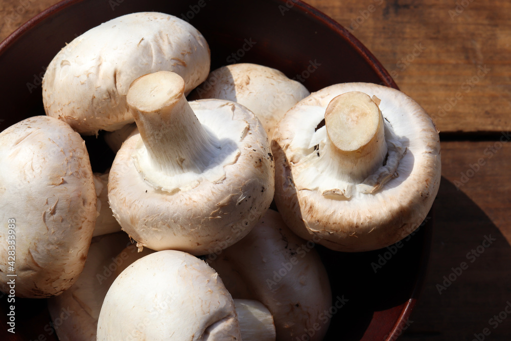 Raw champignon mushrooms in brown ceramic bowl on wooden table background. Top view, copy space. Hard light, shadow	