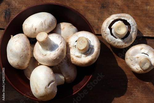 Raw champignon mushrooms in brown ceramic bowl on wooden table background. Top view, copy space. Hard light, shadow 