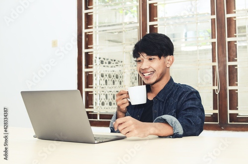 Asian man sits in front of a laptop drinking coffee