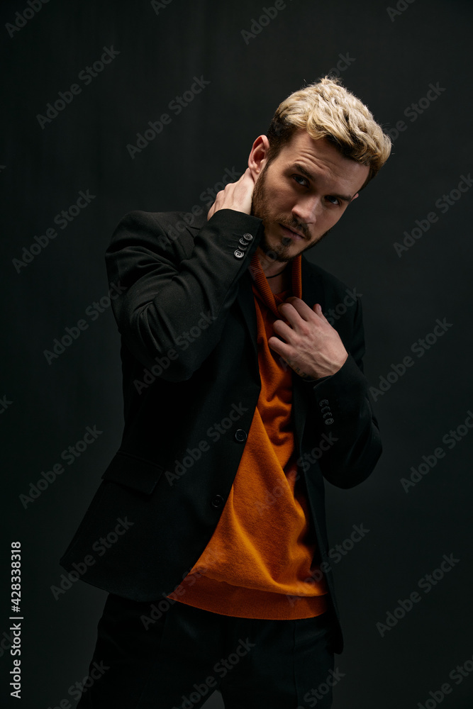 blond guy in an orange sweater and jacket on a dark background holds his hand behind his head blond guy in an orange sweater and jacket on a dark background holds his hand behind his head
