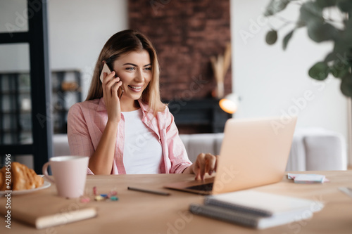 Joyful woman having a phonecall and checking updates on laptop
