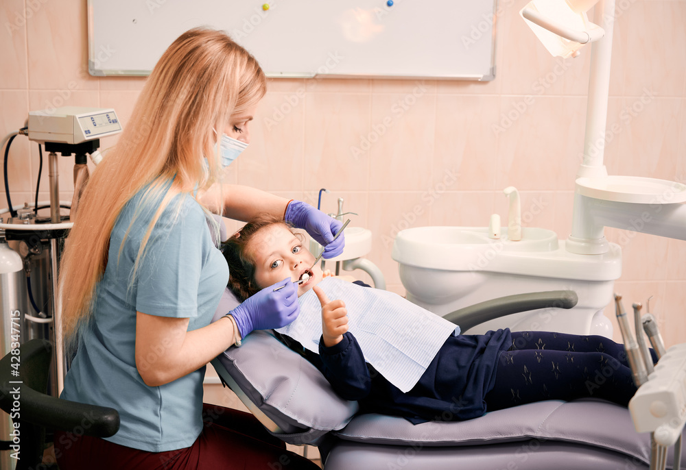 Pediatric dentist checking child teeth with dental explorer and mirror while girl lying in
