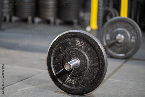 Selective focus at the iron weight plate attached to barbell on the floor inside of fitness gym with blurred background of weight lifting machine for muscle build with copy space. Sport object concept