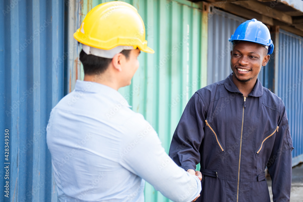 Selective focus at African men worker shake hand with Caucasian freight supervisor while working on site with safety helmet at container logistic import or export dock. Industrial teamwork.