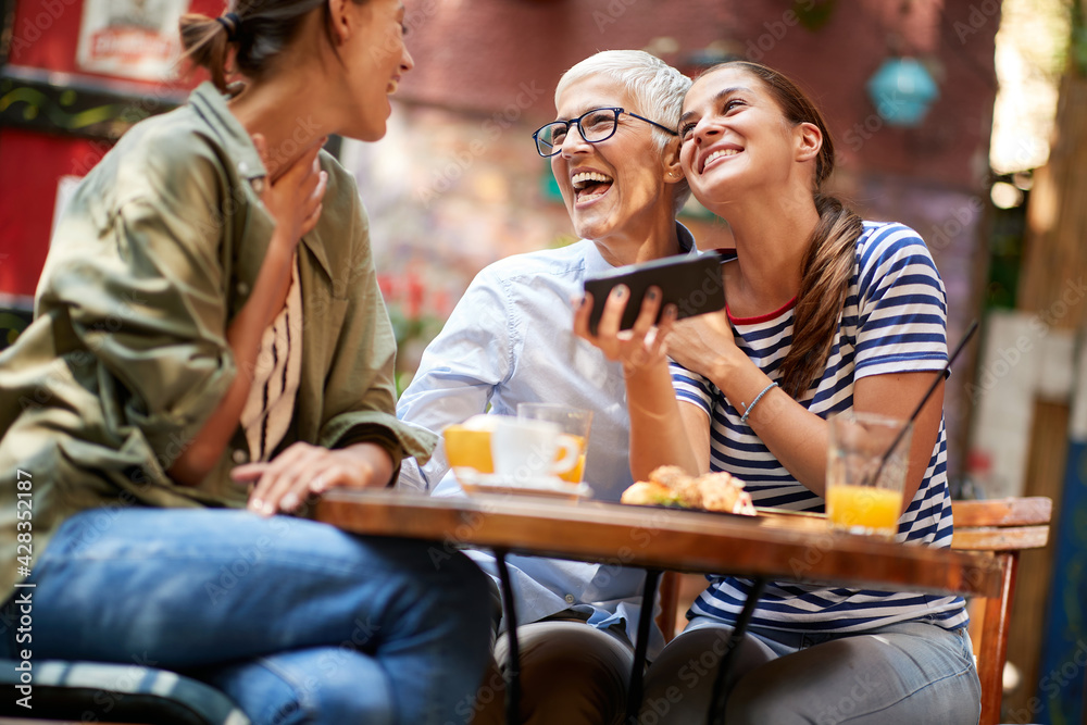 A group of female friends of different generations are laughing at smartphone content they watching while they have a drink in the bar. Leisure, bar, friendship, outdoor - obrazy, fototapety, plakaty 