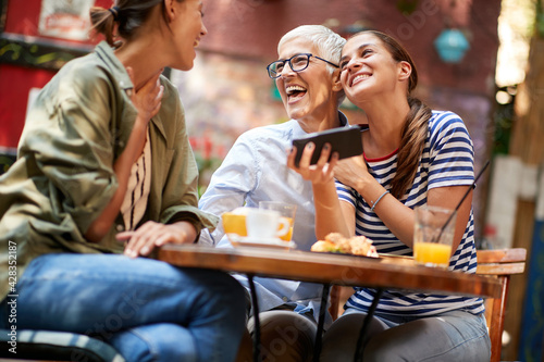 A group of female friends of different generations are laughing at smartphone content they watching while they have a drink in the bar. Leisure, bar, friendship, outdoor