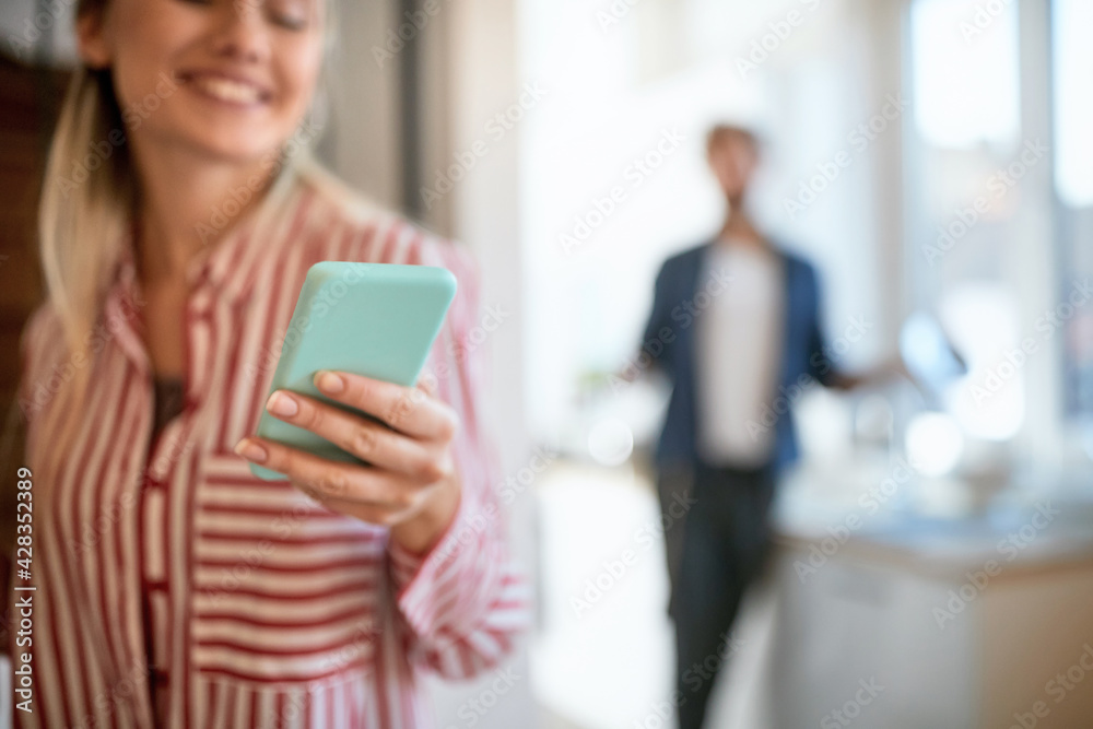young adult blonde holding her cell phone, in focus, with blurry person in the beackground young adult blonde holding her cell phone, in focus, with blurry person in the beackground