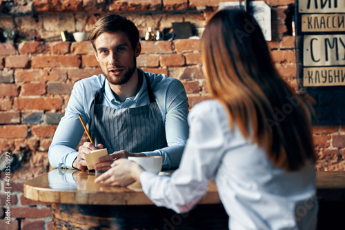 male waiter in gray aprons takes an order and a cup of coffee female client at a table in a cafe