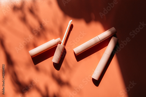 open tube with a brush for liquid lip gloss, liquid lipstick next to closed pink tubes with mascara and eyeliner on a peach background with shadows
