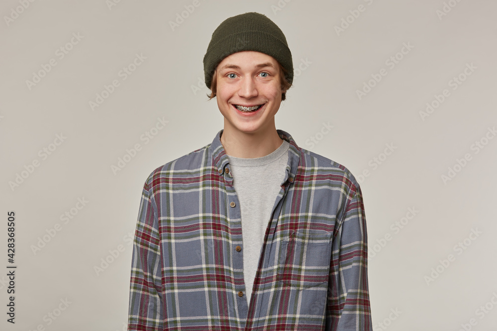 Cheerful guy, happy looking man with blond hair. Wearing checkered shirt and beanie. Has braces. People and emotion concept. Watching positive at the camera isolated over grey background Cheerful guy, happy looking man with blond hair. Wearing checkered shirt and beanie. Has braces. People and emotion concept. Watching positive at the camera isolated over grey background