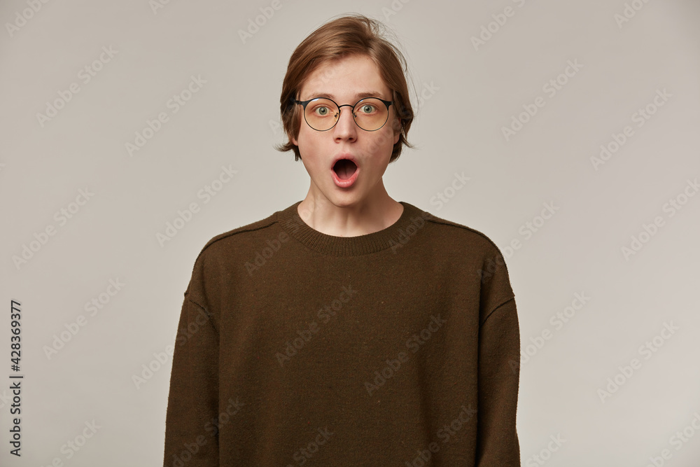 Shocked looking male, handsome guy with blond hair. Wearing brown sweater and glasses. Opens mouth in surprise. People and emotion concept. Watching at the camera isolated over grey background Shocked looking male, handsome guy with blond hair. Wearing brown sweater and glasses. Opens mouth in surprise. People and emotion concept. Watching at the camera isolated over grey background