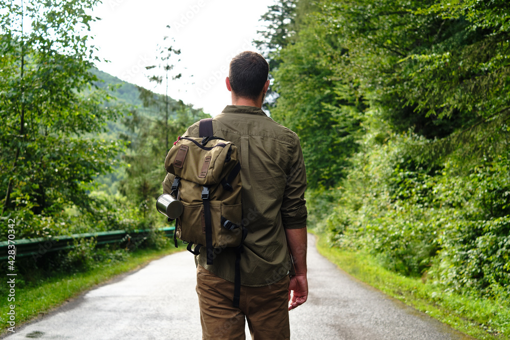 A man with a backpack on an empty road walks forward along the forest. Hike along the hiking trail. Outdoor adventure. Travel and exploration. Healthy lifestyle, active rest