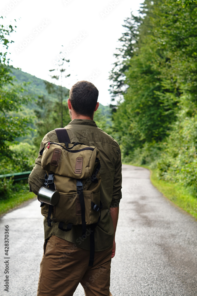 A man with a backpack on an empty road walks forward along the forest. Hike along the hiking trail. Outdoor adventure. Travel and exploration. Healthy lifestyle, active rest