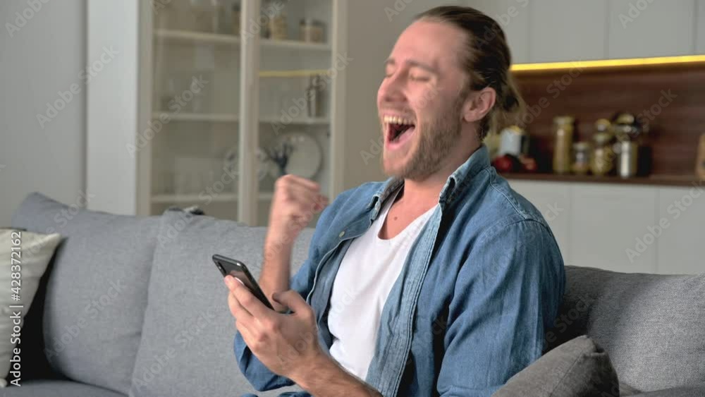 Overjoyed american guy in a stylish denim shirt sits on the sofa in the living room, uses a smartphone, browsing internet, texting with friends, rejoices to win or a good news, smiling happily Overjoyed american guy in a stylish denim shirt sits on the sofa in the living room, uses a smartphone, browsing internet, texting with friends, rejoices to win or a good news, smiling happily