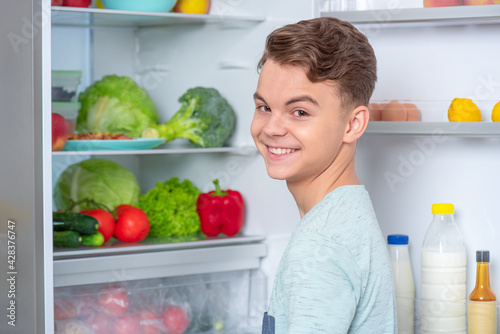 Back view - young teen boy standing near open fridge in kitchen at home. Portrait of pretty child choosing food in refrigerator full of healthy products - rear view