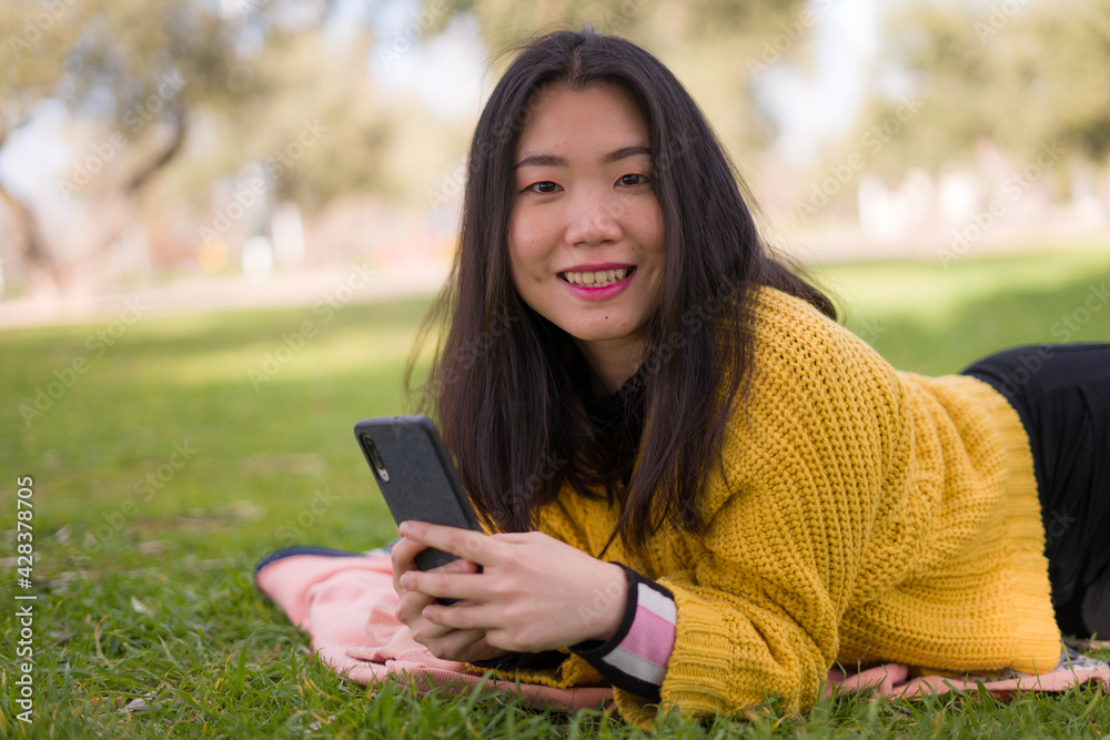 Obraz premium lifestyle portrait of young happy and beautiful Asian Chinese woman in yellow sweater enjoying with mobile phone cheerful at city green park next to tree in Spring