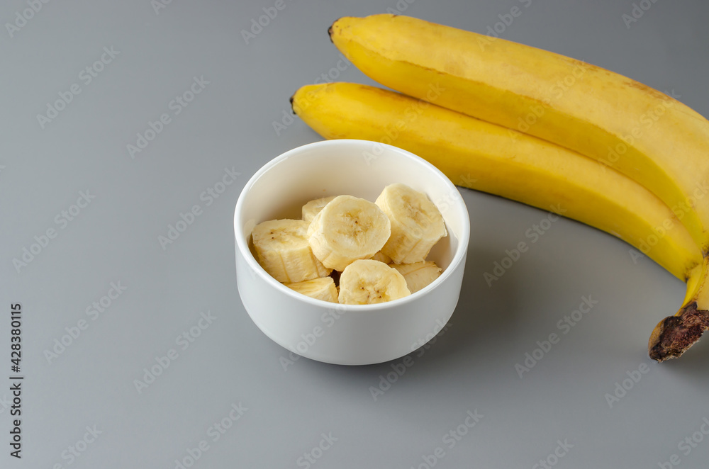 Two bananas and slices in a white plate on a gray background. Trending color of year 2021 Illuminating and Ultimate gray. Side view minimal still life. Two bananas and slices in a white plate on a gray background. Trending color of year 2021 Illuminating and Ultimate gray. Side view minimal still life.