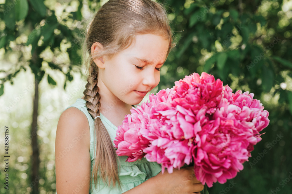 Fototapeta premium portrait of a happy cute little caucasian seven year old kid girl, holds in hands and smell and enjoying a bouquet of pink peony flowers in full bloom on the background of nature