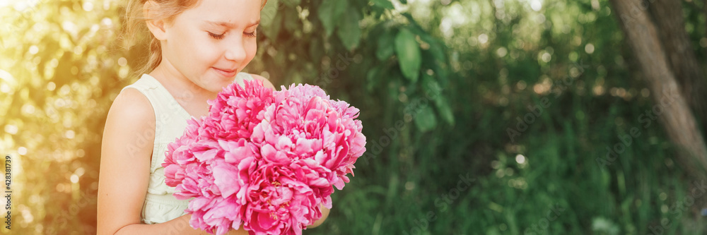 portrait of a happy cute little caucasian seven year old kid girl, holds in hands and smell and enjoying a bouquet of pink peony flowers in full bloom on the background of nature. banner. flare portrait of a happy cute little caucasian seven year old kid girl, holds in hands and smell and enjoying a bouquet of pink peony flowers in full bloom on the background of nature. banner. flare