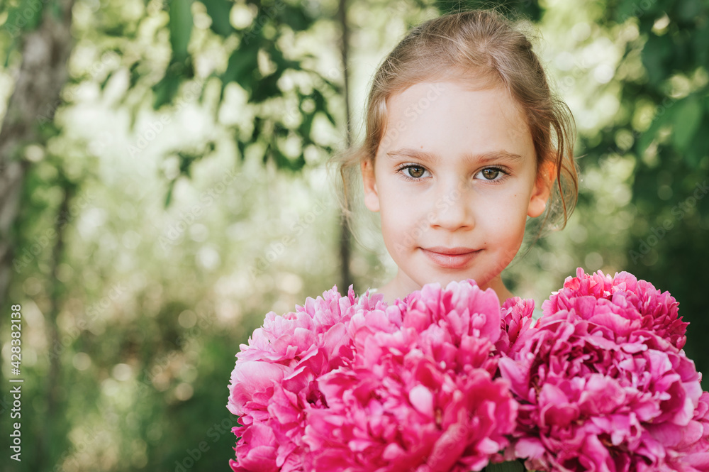 Fototapeta premium portrait of a happy cute little caucasian seven year old kid girl, holds in hands a bouquet of pink peony flowers in full bloom on the green background of nature