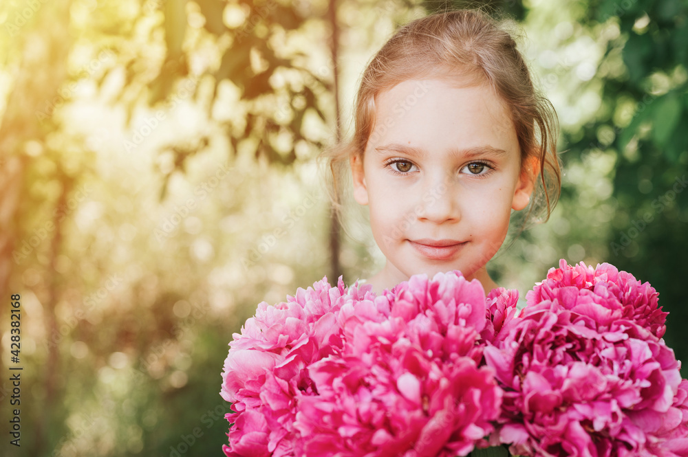 Naklejka premium portrait of a happy cute little caucasian seven year old kid girl, holds in hands a bouquet of pink peony flowers in full bloom on the green background of nature. flare