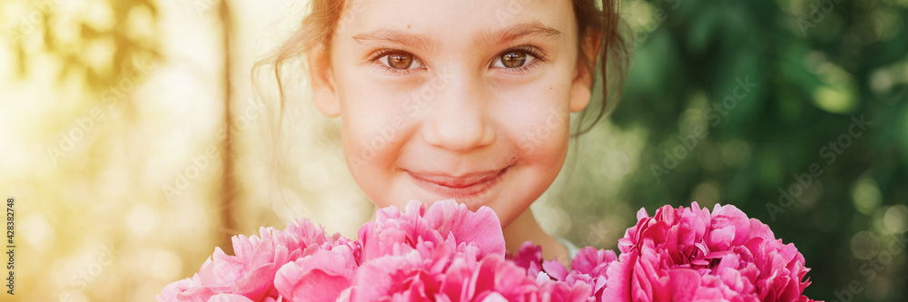portrait of a happy cute little caucasian seven year old kid girl, holds in hands a bouquet of pink peony flowers in full bloom on the green background of nature. banner. flare