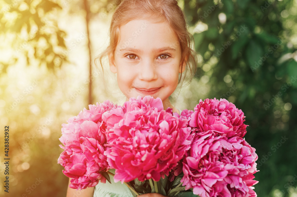 Fototapeta premium portrait of a happy cute little caucasian seven year old kid girl, holds in hands a bouquet of pink peony flowers in full bloom on the green background of nature