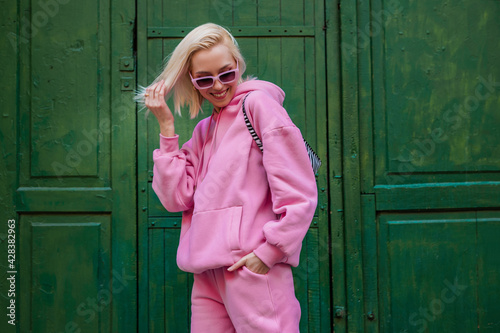 Fototapeta Naklejka Na Ścianę i Meble -  Spring fashion concept: happy smiling fashionable woman wearing trendy pink sport chic style outfit posing in street on green background. Sunglasses, pink hoodie, trousers. Copy, empty space for text