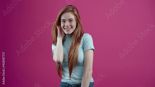 The red-haired teenager listens to the new rumor that surprises her and makes her smile, in a casual blue T-shirt, isolated on a pink background. The concept of people's lifestyle photo