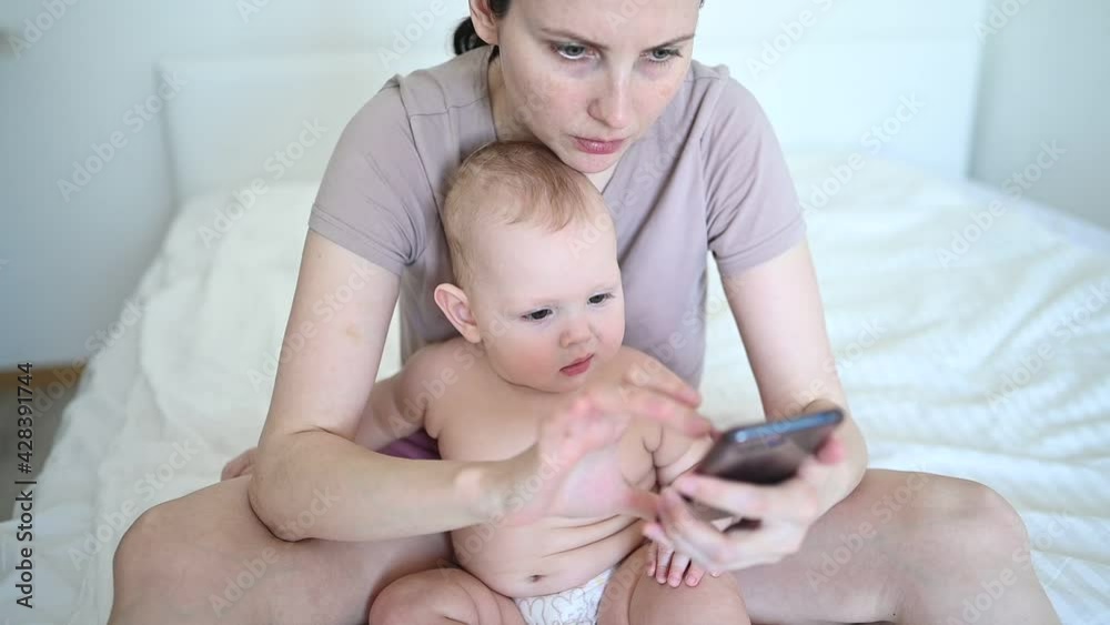 Little baby toddler boy interested in watching mother who works with smartphone phone. Child and modern technology. Happy family relaxing in bedroom early morning Little baby toddler boy interested in watching mother who works with smartphone phone. Child and modern technology. Happy family relaxing in bedroom early morning