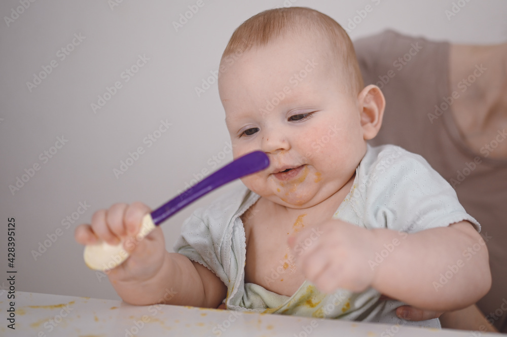 Little newborn funny baby boy learning to eat vegetable or fruit puree from glass jar with spoon. Young mother helping little son eat first food Little newborn funny baby boy learning to eat vegetable or fruit puree from glass jar with spoon. Young mother helping little son eat first food