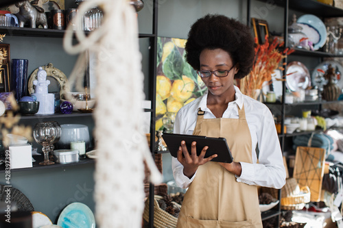 Pretty afro american woman in eyegasses using digital tablet for making inventory at decor shop. Attractive saleswoman in apron standing with modern gadget in hands. photo