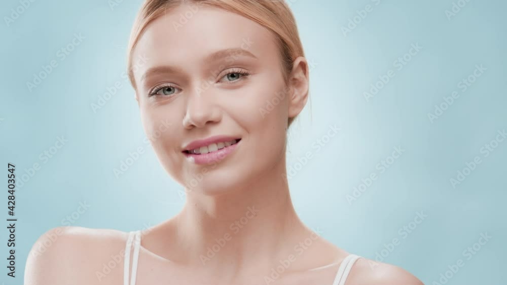 Young slim pretty blonde Caucasian woman with ponytail in white crop top turns her head to the camera touching her shoulder and smiling wide against blue ripple background | Skincare commercial Young slim pretty blonde Caucasian woman with ponytail in white crop top turns her head to the camera touching her shoulder and smiling wide against blue ripple background | Skincare commercial