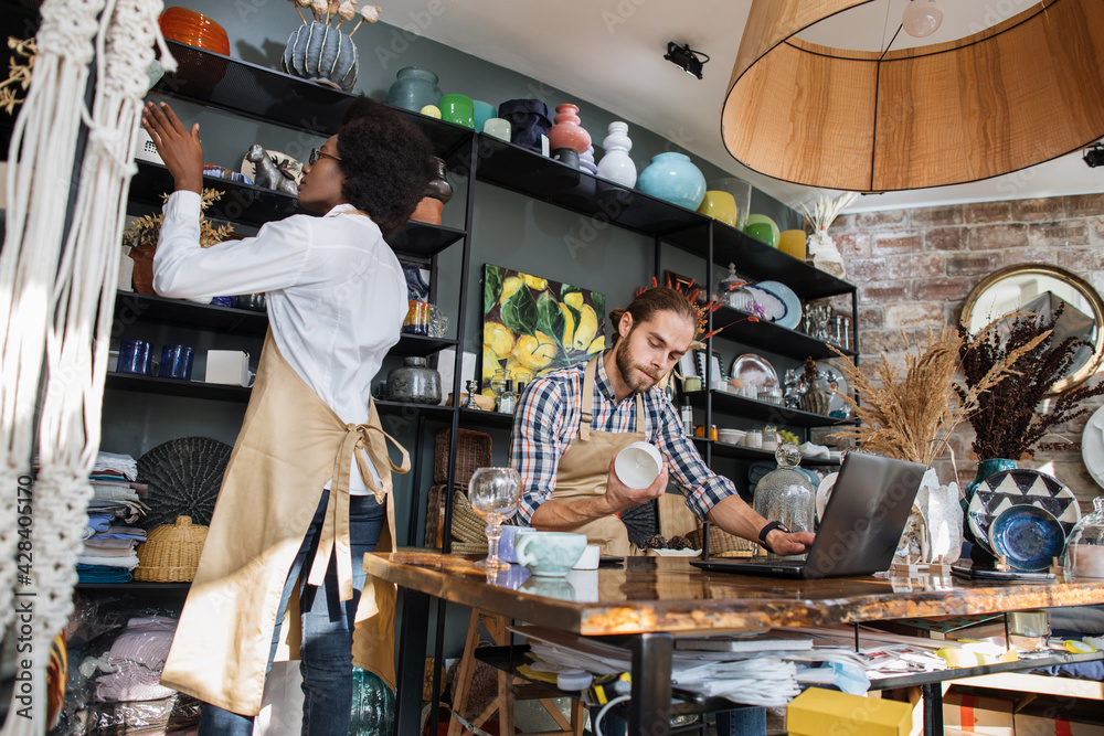 Two multicultural workers in uniform doing inventory together at decor shop. Bearded caucasian man using laptop while african woman counting goods.