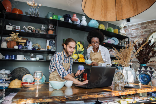 Two multicultural people working on modern laptop during inventory process at decor store. Young caucasian man and african woman rechecking all assortment at shop. photo