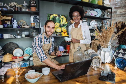 Two smiling multiethnic people working on modern laptop during inventory process at decor store. Young caucasian man and african woman rechecking all assortment at shop. photo