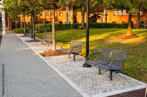 Empty Stylish steel benches along a paved path in a public park surrounded by brick buildings at sunset