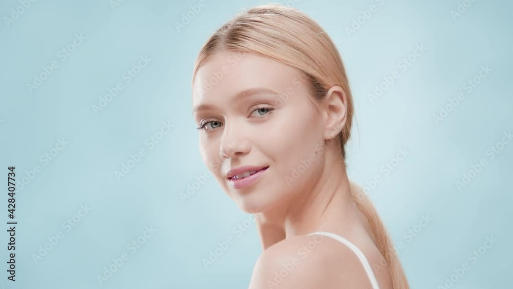 Young fit good-looking Caucasian woman with long golden hair in ponytail in white top touches her jawline turning her body and looking at the camera against blue ripple background | Skincare concept Young fit good-looking Caucasian woman with long golden hair in ponytail in white top touches her jawline turning her body and looking at the camera against blue ripple background | Skincare concept