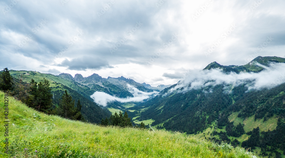 Praderas verdes, bosques de coníferas, nubes bajas y altas de lluvia penetrando por un valle lateral del gran valle de Arán, en los Pirineos españoles