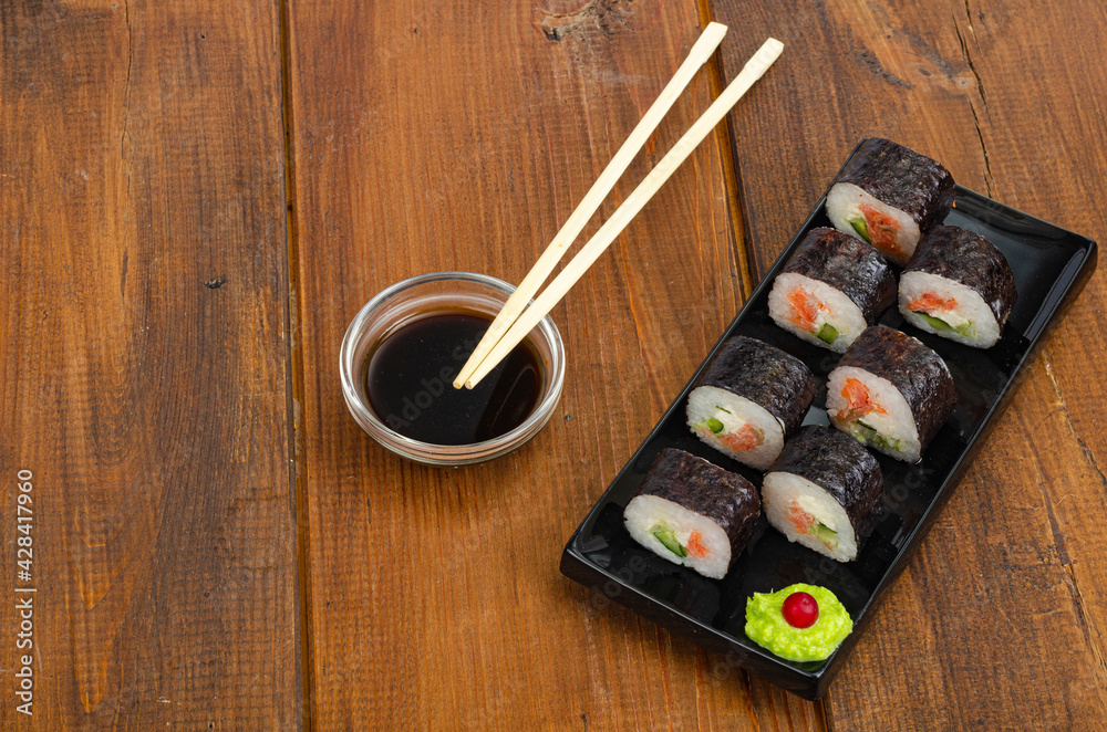 Black plate with rolls with salmon on wooden table. Green wasabi and soy sauce. Black plate with rolls with salmon on wooden table. Green wasabi and soy sauce.