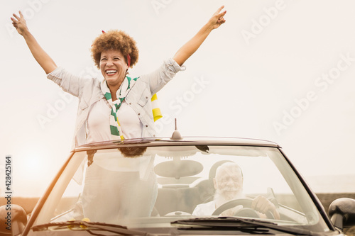 Happy senior couple having fun in summer vacation with cabriolet car - Elderly multiracial people inside convertible car