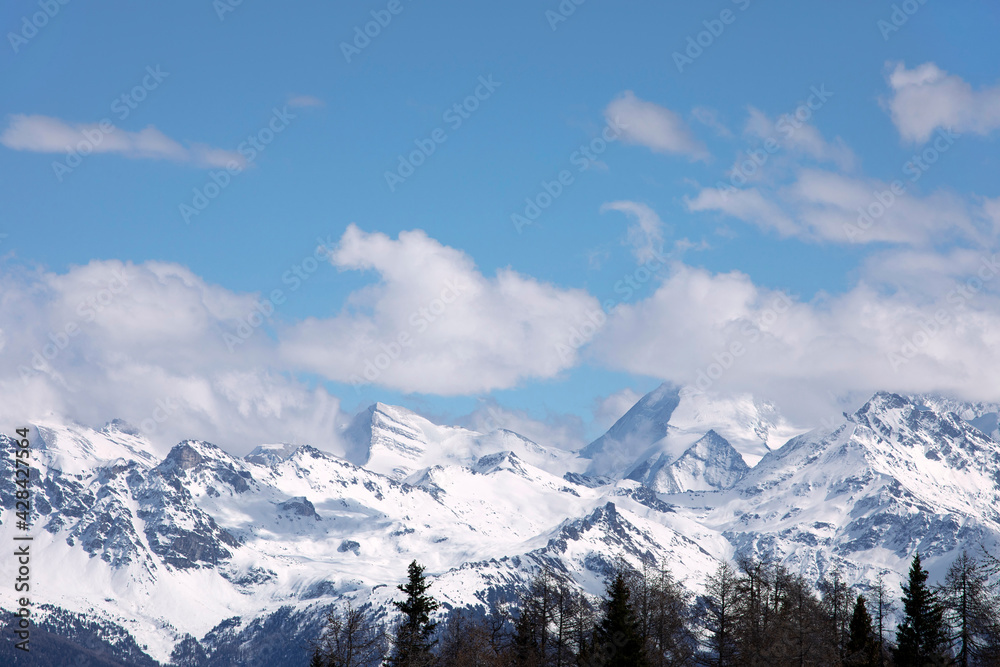 Alpine landscape with tops of fir trees. Winter landscape, with snowcapped distant mountains, blue sky with clouds, sunny day. Snowy mountain peaks in Swiss alps, Wallis canton.