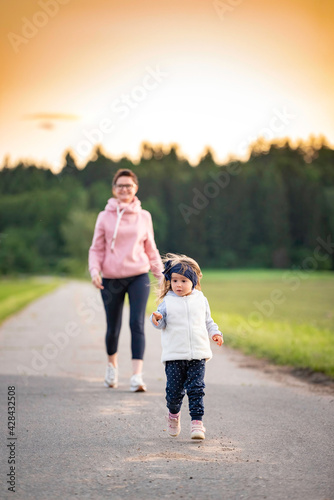 Mother and child walking on countryside road between agricultural fields towards vilage from forest.