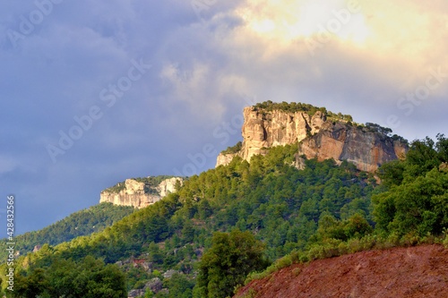 Orange massive rock on green mountainside covered with trees, climbing sector. Blue sky with white clouds. Beautiful nature, mountains. Siurana, Catalonia, Spain.