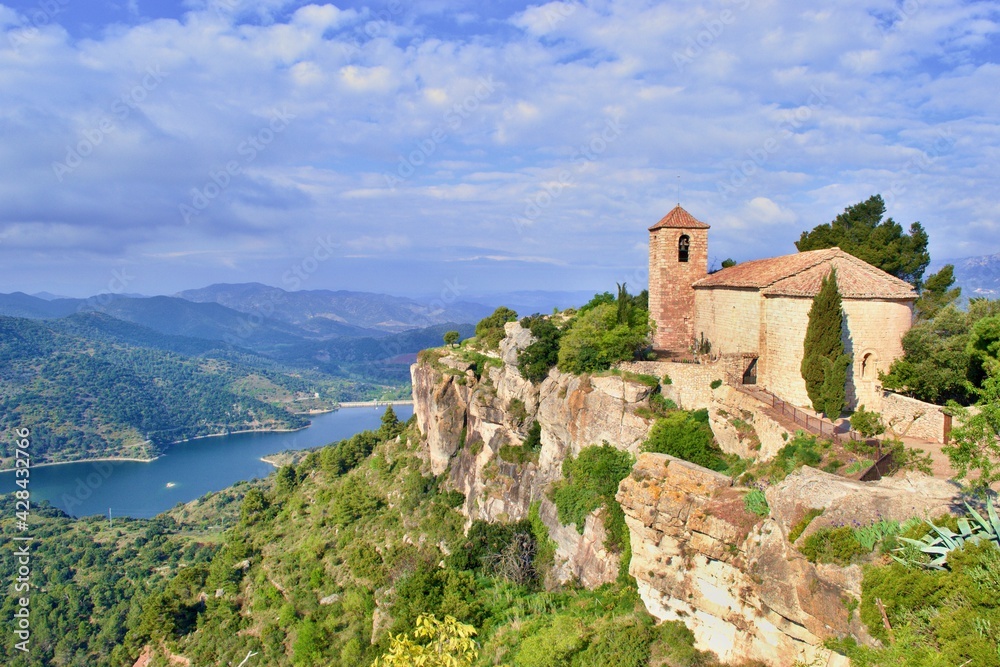 Romanesque church of Santa Maria de Siurana on top of rock, view into valley, mountains on the background. Blue sky with white clouds.  Siurana, Catalonia, Spain.