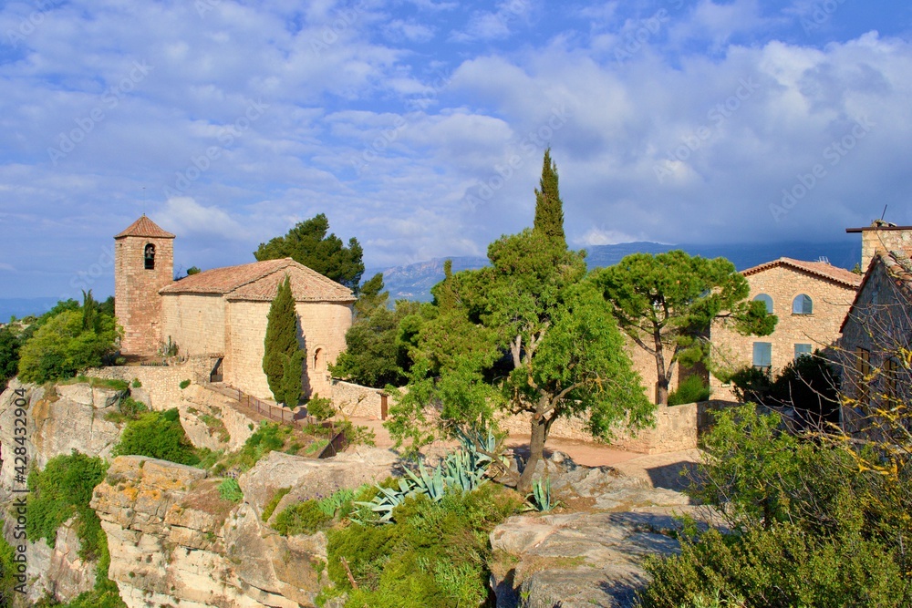Village with romanesque church of Santa Maria de Siurana on top of rock, view into valley, mountains on the background. Blue sky with white clouds.  Siurana, Catalonia, Spain.
